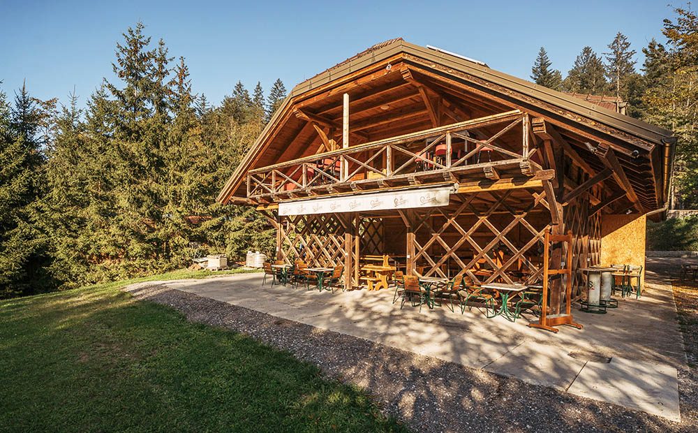 Zunanji pokrit prostor Toplar spodaj, obdan z zeleno travo in smrekami.